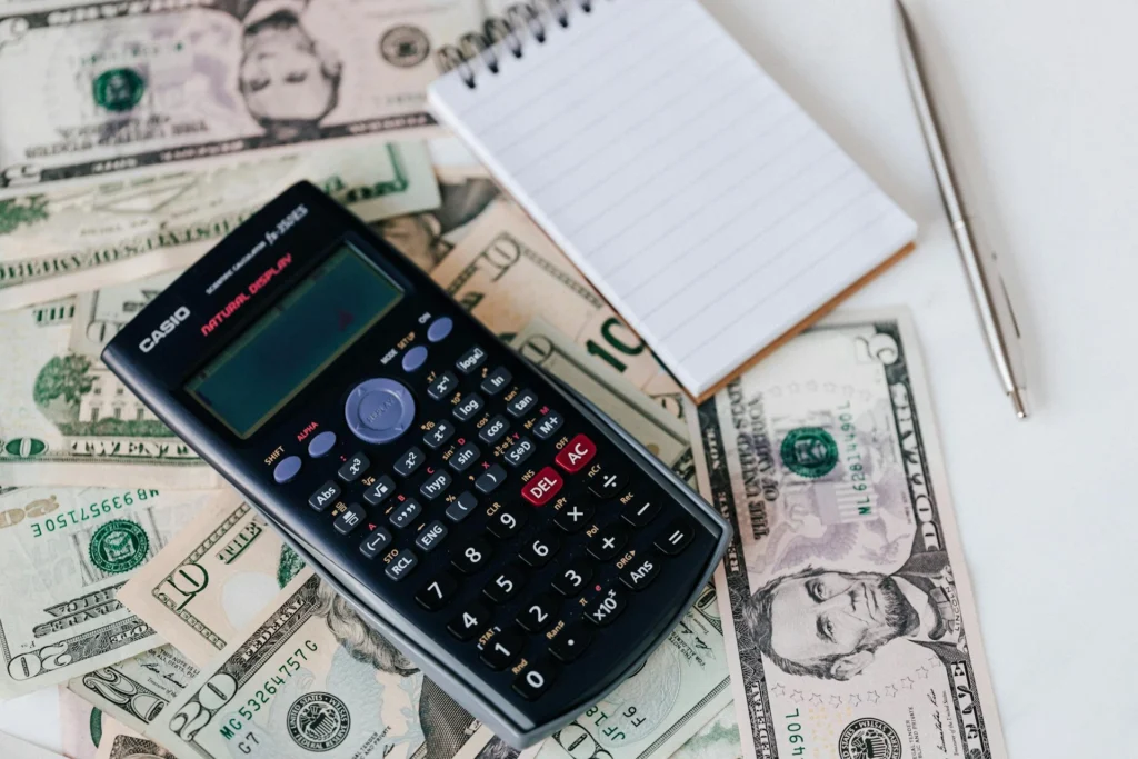 A calculator, pen and notepad on a table next to a pile of money