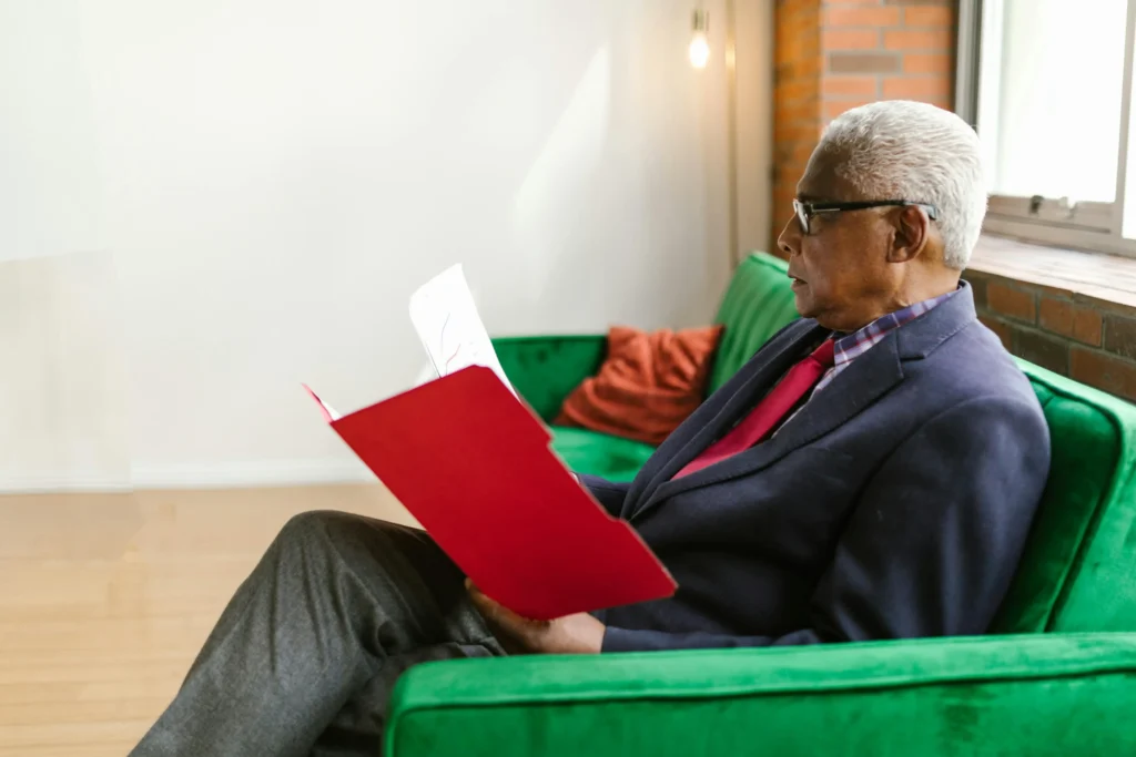An elderly man in blue suit sitting on the couch while looking at the red folder.