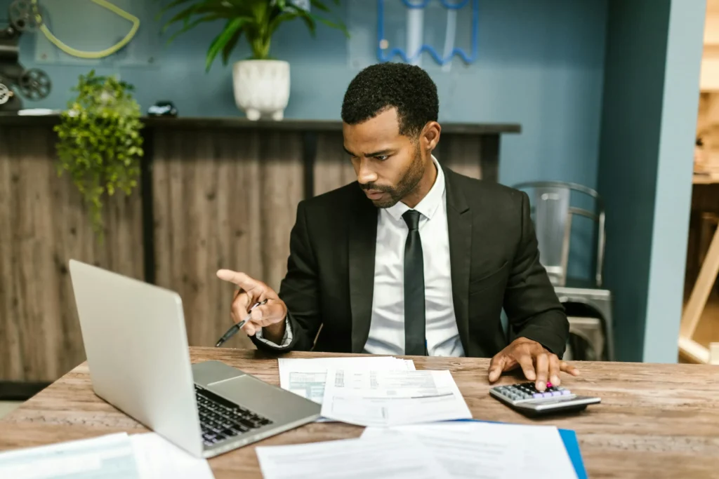 Professional man in a suit reviewing financial documents at a desk with a laptop and calculator, appearing focused and analytical.